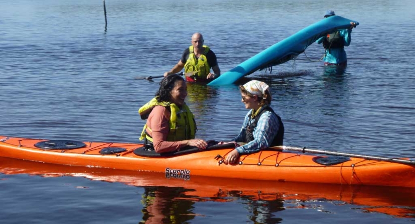 In the foreground, a person sitting in an orange kayak listens to an instructor who is standing in waist-deep water. Behind them, another person wades through the water while another braces a blue kayak on their shoulders. 